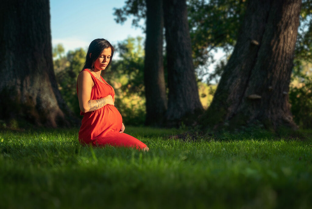 Gravid kvinna sitter på knä i röd klänning på gräset Sågudden Arvika