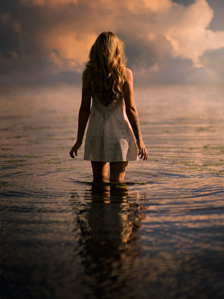 kvinna i vit klänning står i sjön vid solnedgången