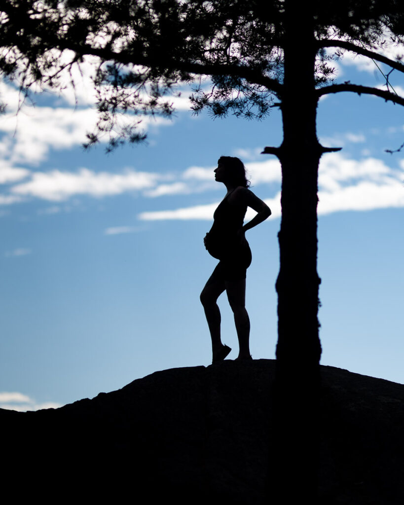Silhuett av gravid kvinna på sten vid träd mot blå himmel
