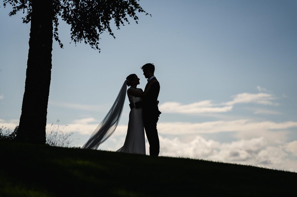 Brudpar håller om varandra stående på en kulle vid ett träd med himmel som bakgrund.
