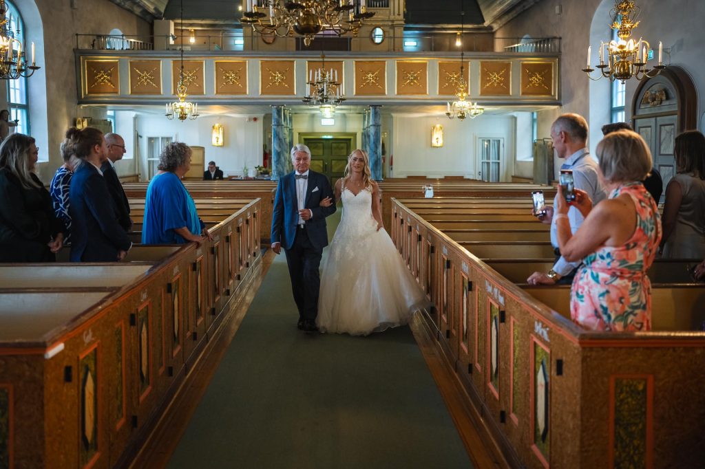 Bruden kommer arm I arm med sin far nedför altargången i Mikaelikyrkan i Arvika.