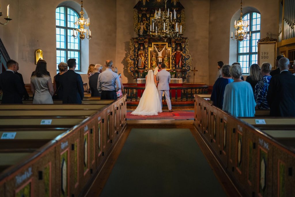 Brudparet står hand i hand framför prästen och altaret i Mikaelikrykan i Arvika.