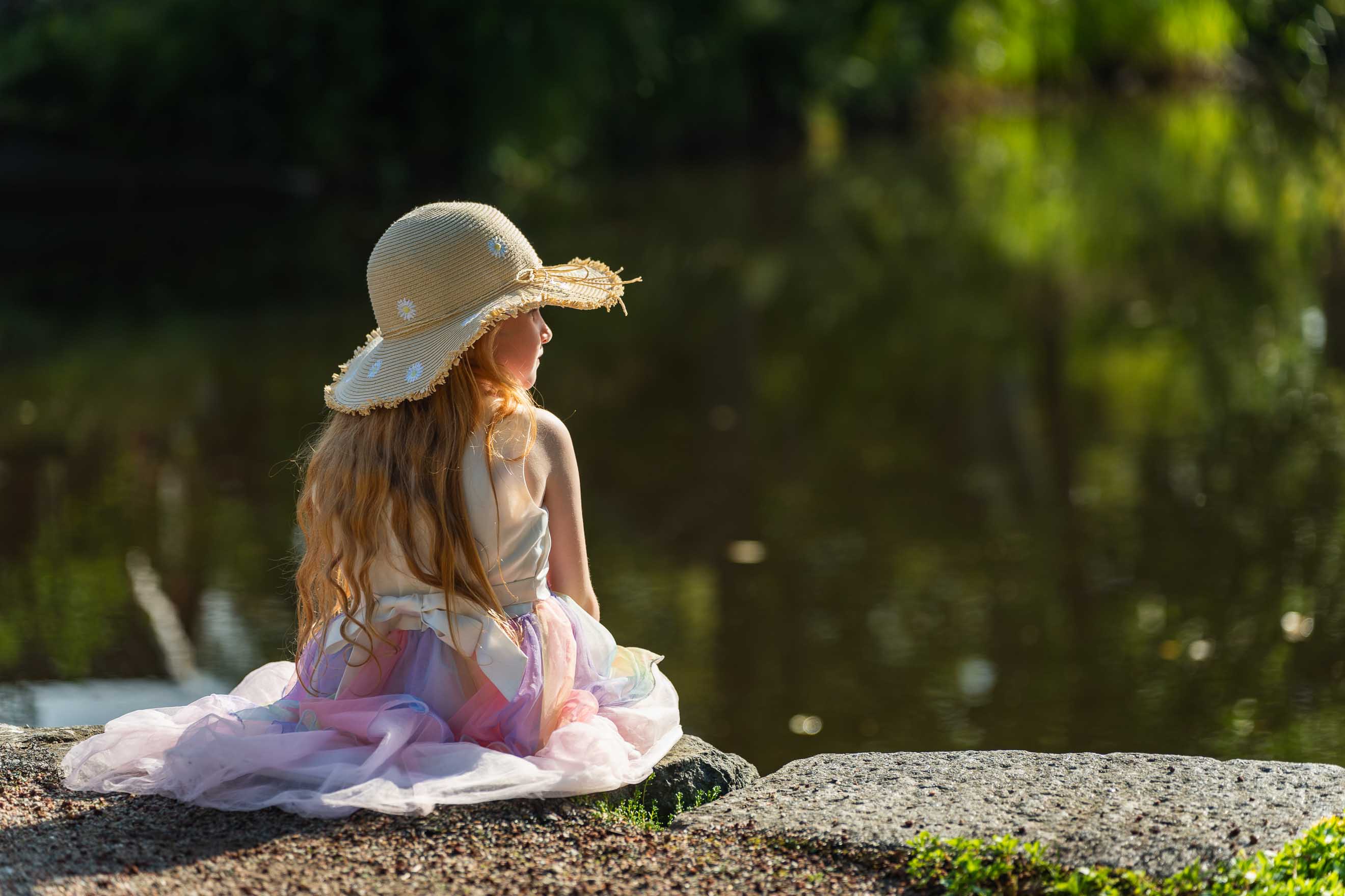 Liten flicka i stråhatt sitter vid sjön.