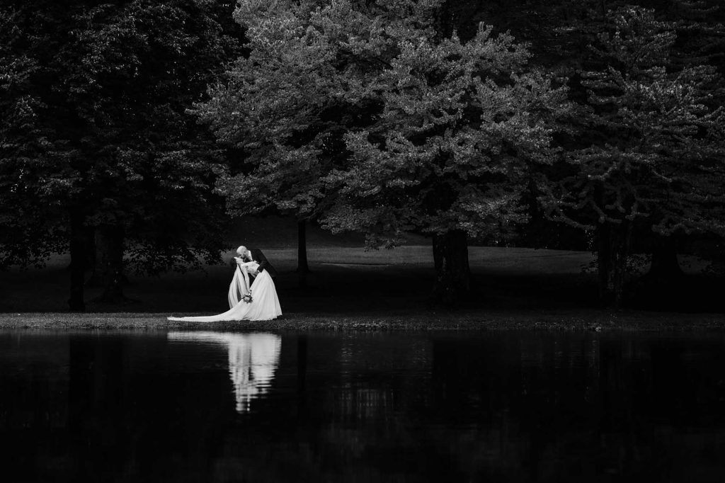 Brudgummen håller bakom ryggen på bruden och böjer henne ned för en kyss vid en damm.