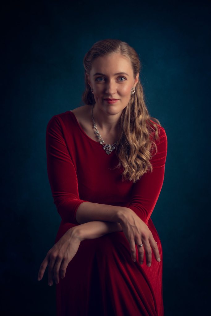 Blond kvinna i röd klänning sitter på pall med ben och armar i kors.