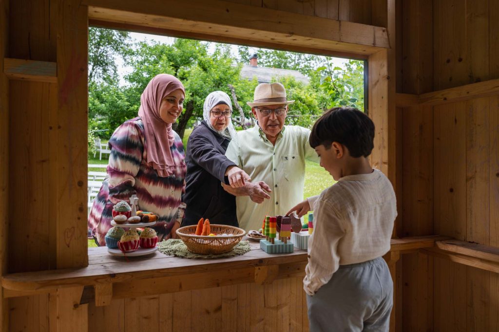 Invandrarfamilj i lekstuga/café, barn serverar kaffe på Rackstadsmuseet Arvika till morföräldrar.