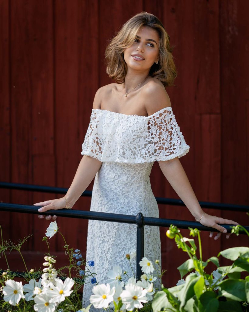 Ung kvinna i vit klänning står vid röd husvägg med vita blommor i förgrunden.