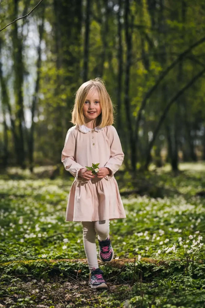 En leende flicka med en vitsippa i handen under en barnfotografering utomhus i en lummig skog i Kil.