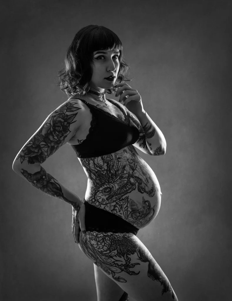 Svartvit studiobild på en gravid kvinna med konstnärliga tatueringar, fotograferad med dramatiskt ljus i en fotostudio hos PixPep i Arvika.