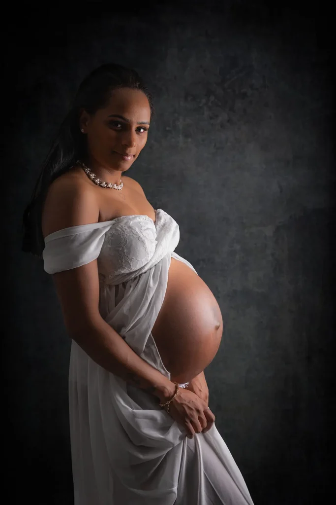 Ett vackert och tidlöst gravidporträtt i studio på en kvinna i vit klänning mot mörk bakgrund, fotad hos PixPep i Arvika.