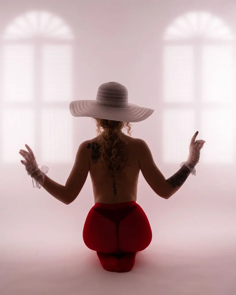 Ett konstnärligt och grafiskt boudoir-porträtt på en kvinna i hatt och röda detaljer mot en ljus bakgrund med fönsterskuggor, fotat i vår studio i Arvika.