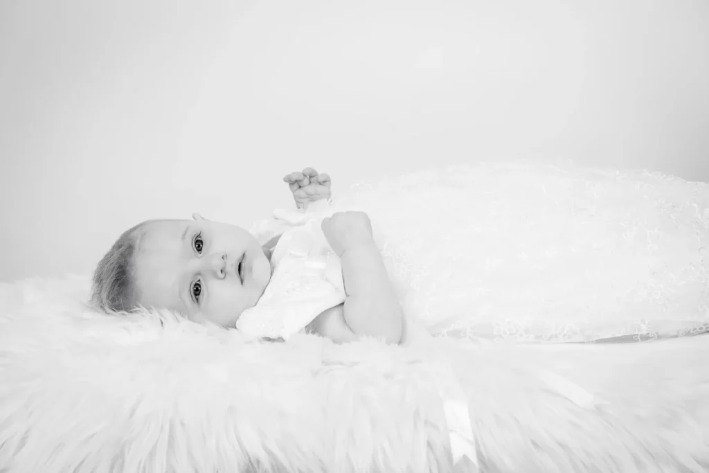 Mjukt svartvitt barnporträtt av bebis i spetsklänning (dopklänning) liggande på fuskpälshår, fotograferat med naturligt studioljus i en studio i Arvika med ljus bakgrund.