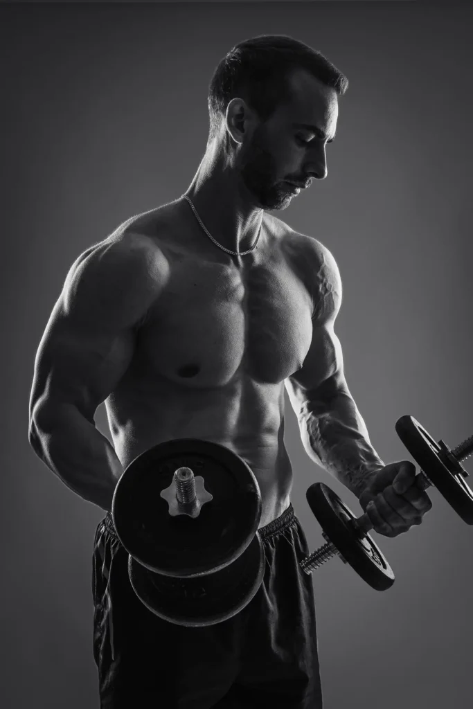 Dramatiskt svartvitt fitnessporträtt av en muskulös man med bar överkropp som lyfter hantlar, fotograferat med kantljus i en studio i Arvika med mörk bakgrund.