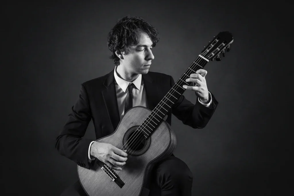 Stämningsfullt svartvitt studioporträtt av musiker i kostym som spelar klassisk gitarr, fotograferat med dramatiskt kantljus i en studio i Arvika med mörk bakgrund.