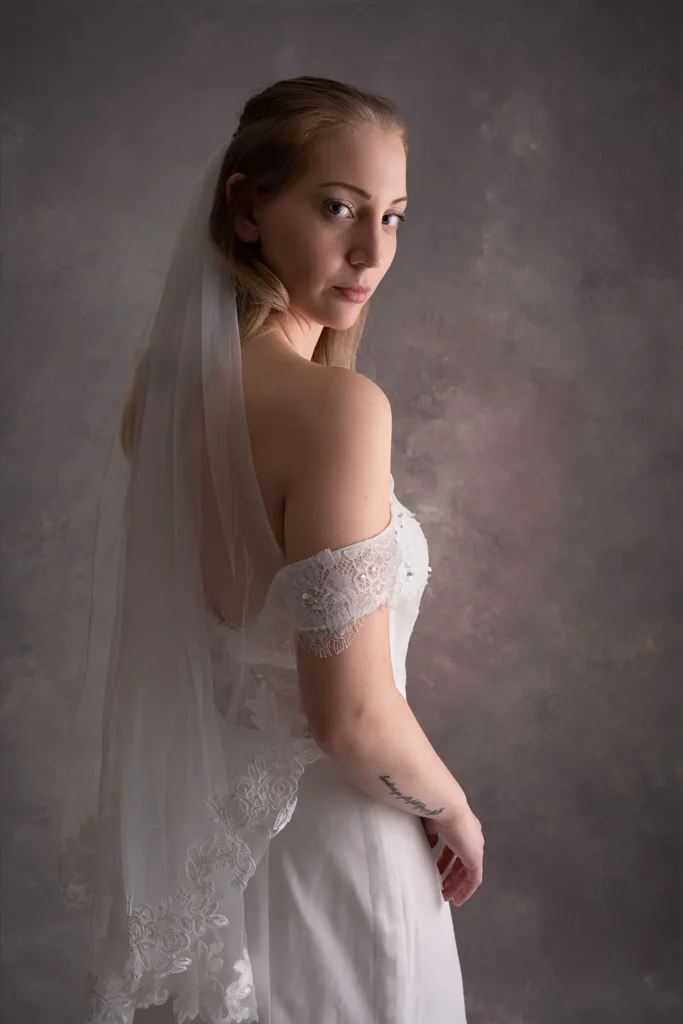 Professionell studiobild av brud sedd bakifrån, med blicken över axeln, bärandes en off-shoulder brudklänning med spetsdetaljer och en slöja mot en grå, texturerad bakgrund.