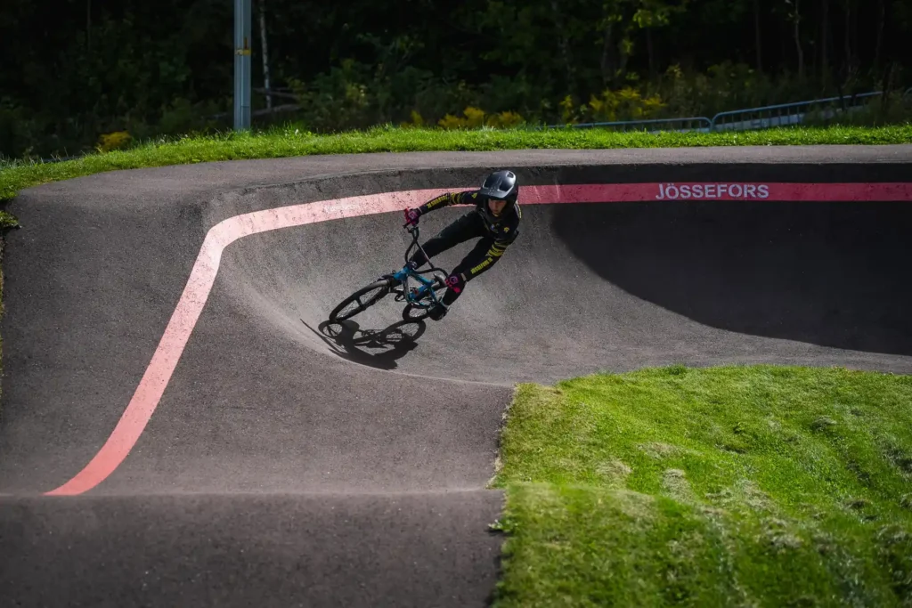 En cyklist lutar sig djupt i en kurva på den asfalterade pumptrackbanan i Jössefors under en solig dag.