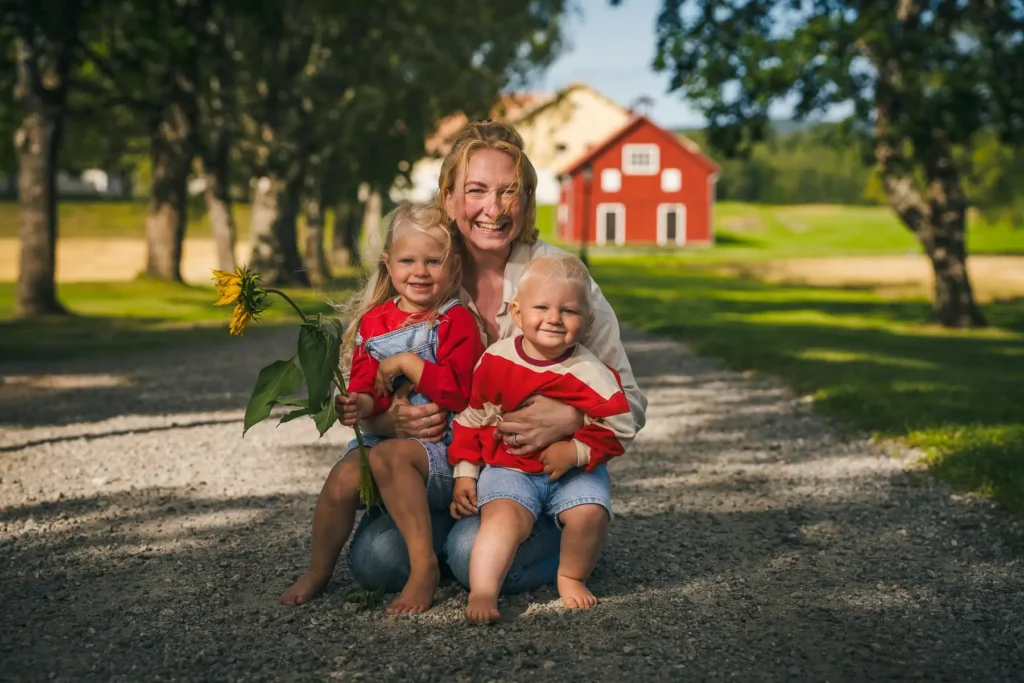 En glad kvinna som kramar om två barn på en grusgång vid Ruds Säteri i Arvika, med en röd lada i bakgrunden och en solros i förgrunden.