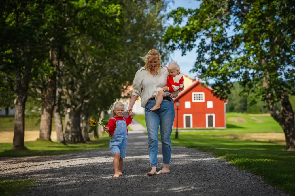 En barfota flicka i hängselbyxor håller en solros och går bredvid sin mamma på en grusallé vid Ruds säteri i Arvika.