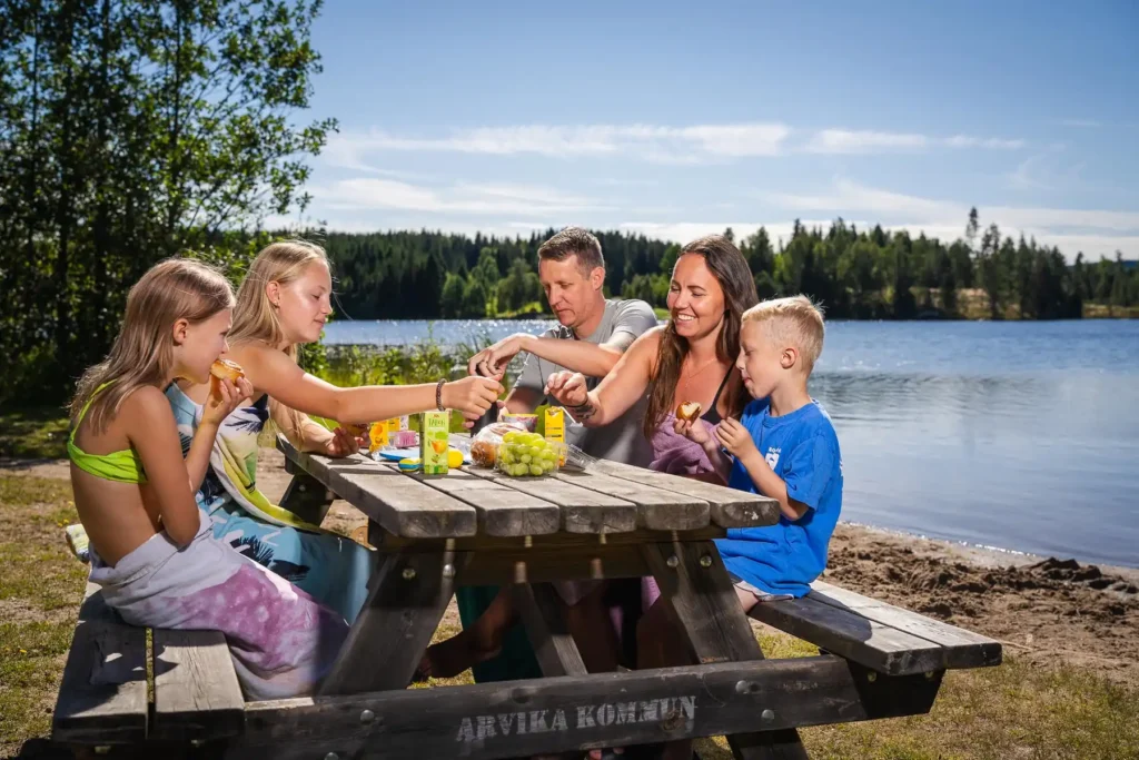 En kvinna och två barn sitter på en filt och fikar vid strandkanten till Kolsjön i Arvika.