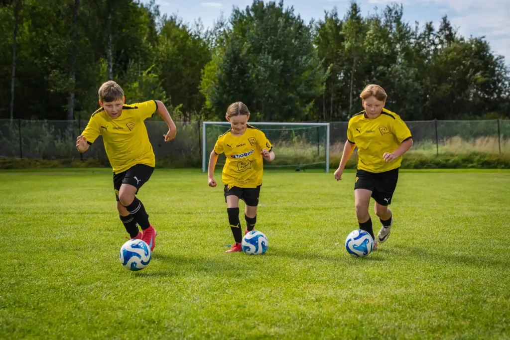 Tre barn i gula lagtröjor driver bollen framåt under en fotbollsträning på Forshälla idrottsplats i Jössefors.