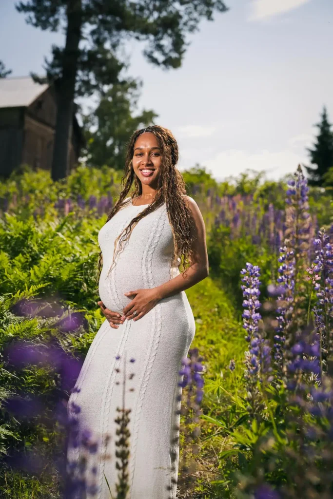 En strålande gravid kvinna i vit klänning står i ett fält av blommande lupiner i Tasebo, Arvika, fotograferad av en gravidfotograf verksam i Värmland.
