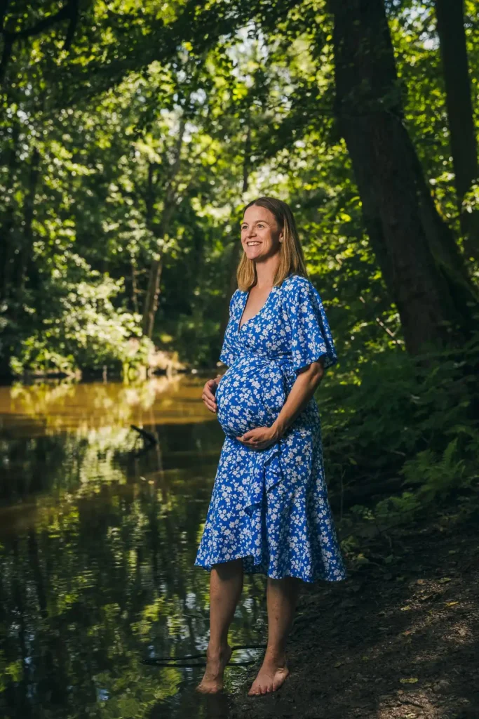 En leende gravid kvinna i en blå blommig klänning står barfota vid ett vattendrag i en lummig skogsmiljö i Karlstad, fotograferad av PixPep Gravidfotograf i Värmland.