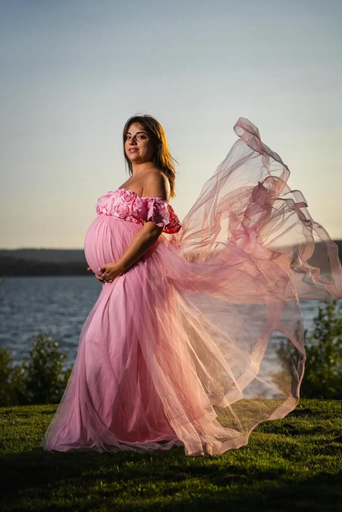En gravid kvinna i en böljande rosa tyllklänning vid Vänerns strand på Duse Udde i Säffle, fotograferad av en professionell fotograf.
