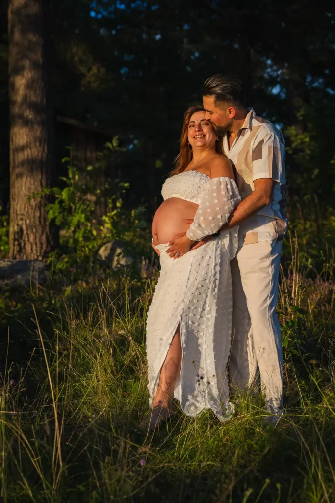 Ett lyckligt par under en gravidfotografering i en solig skogsmiljö i Säffle, där pappan pussar mamman på pannan i det mjuka motljuset.