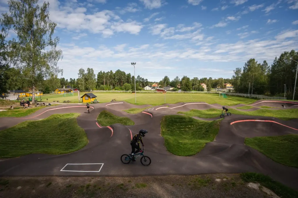 En stor översiktsbild över Jössefors asfalterade pumptrackbana i Arvika, med flera cyklister och fotbollsplanen i bakgrunden.